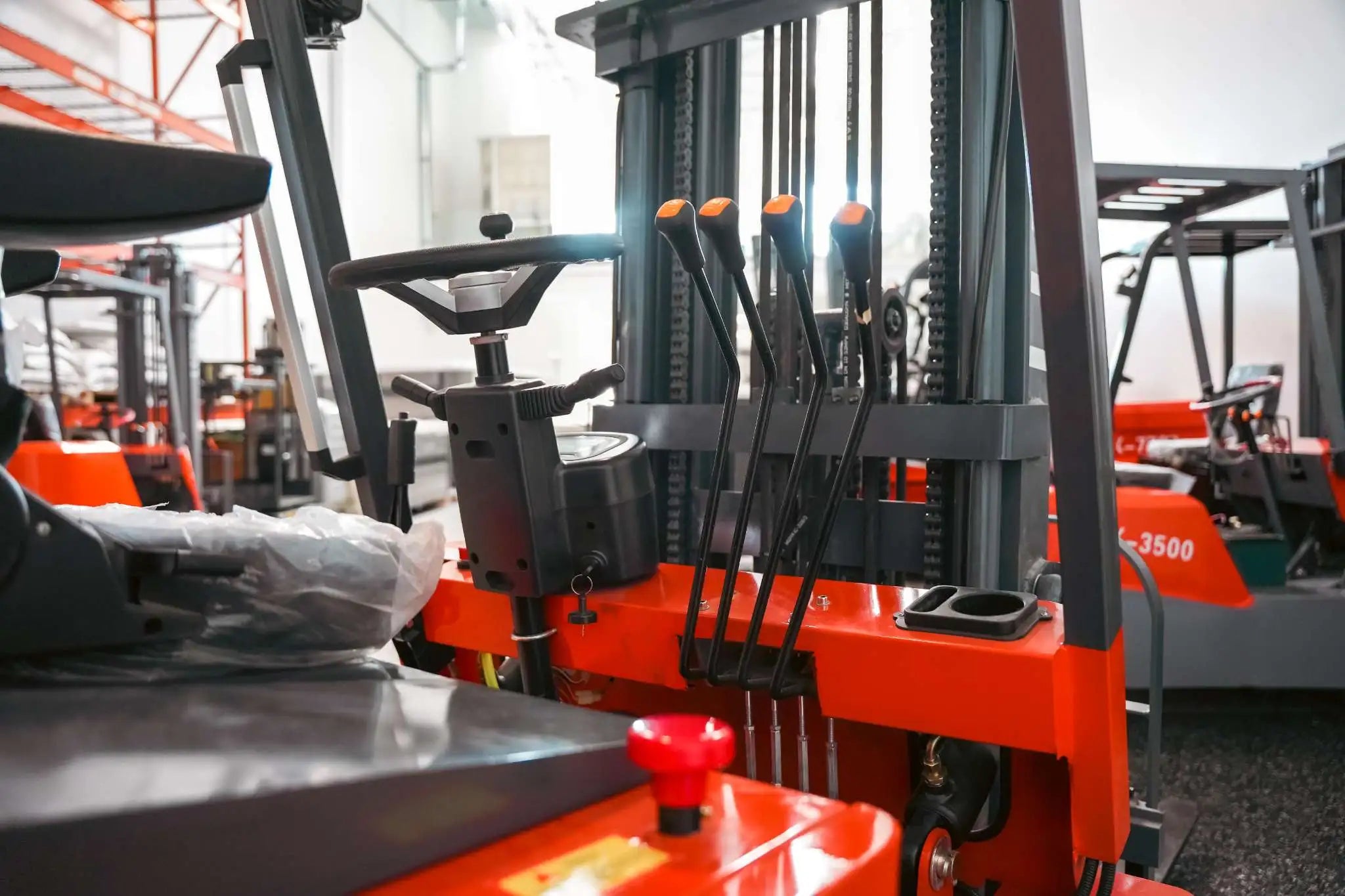 LUXX LX-4400 Compact forklift cabin detail showing orange-tipped hydraulic levers, red emergency stop button, steering column, and mast chains in a large warehouse facility.