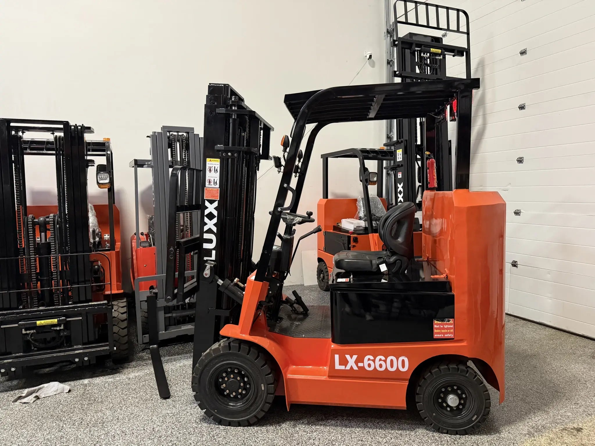 LUXX LX-6600 Compact orange forklift in a full showroom floor view, with multiple LUXX units and masts stacked in the background, showing compact size in context.
