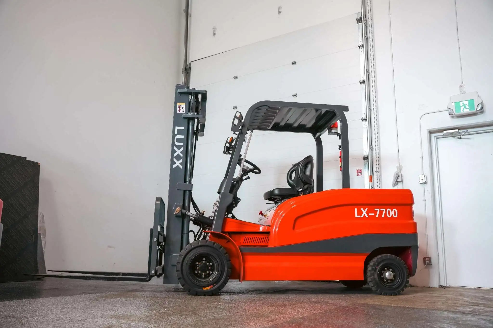LUXX LX-7700 red forklift with grey mast, overhead guard, and solid tyres, photographed from the side inside an industrial warehouse facility.