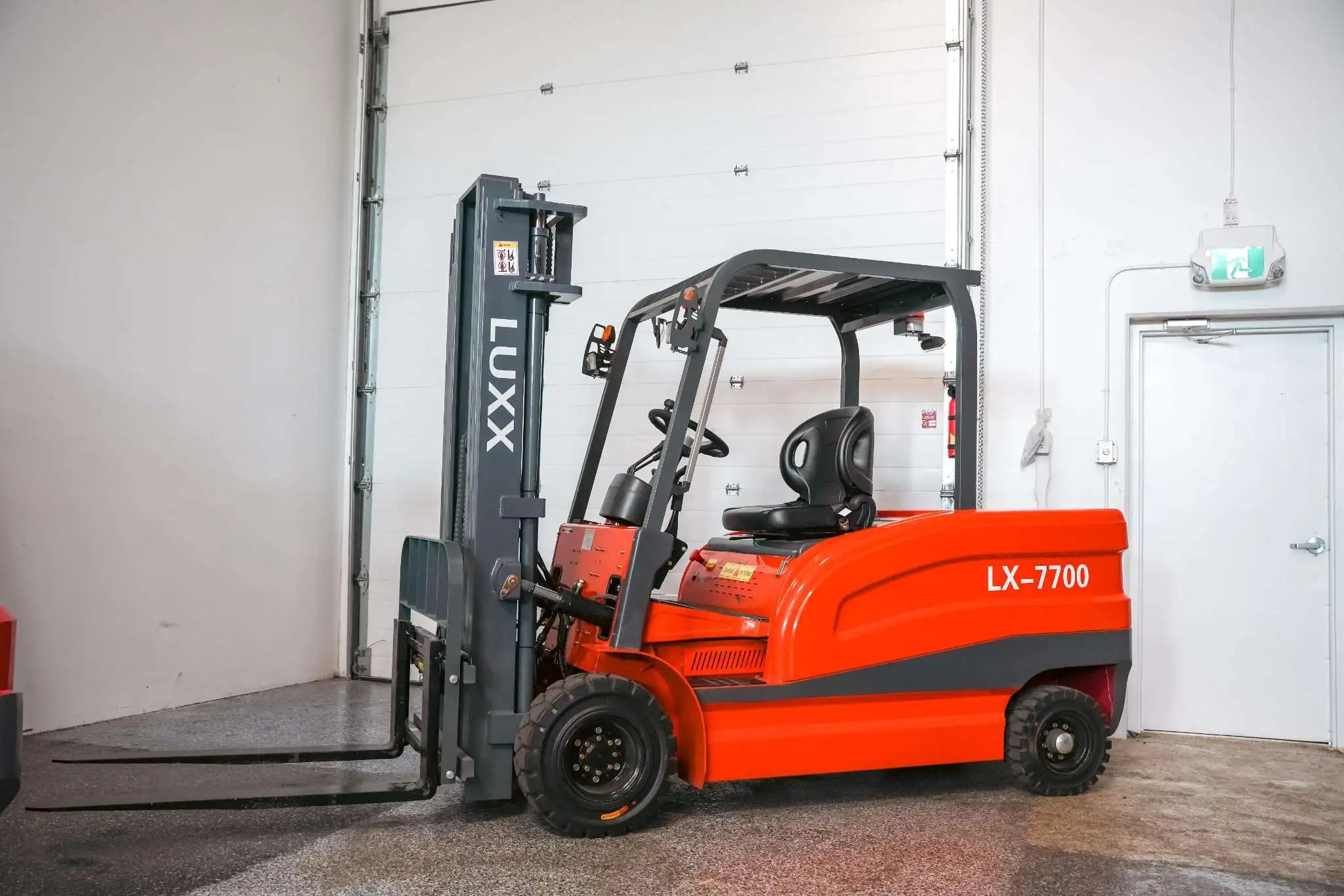 LUXX LX-7700 red forklift viewed from a three-quarter front angle, displaying the full body, grey mast with forks lowered, and open operator cabin in a warehouse