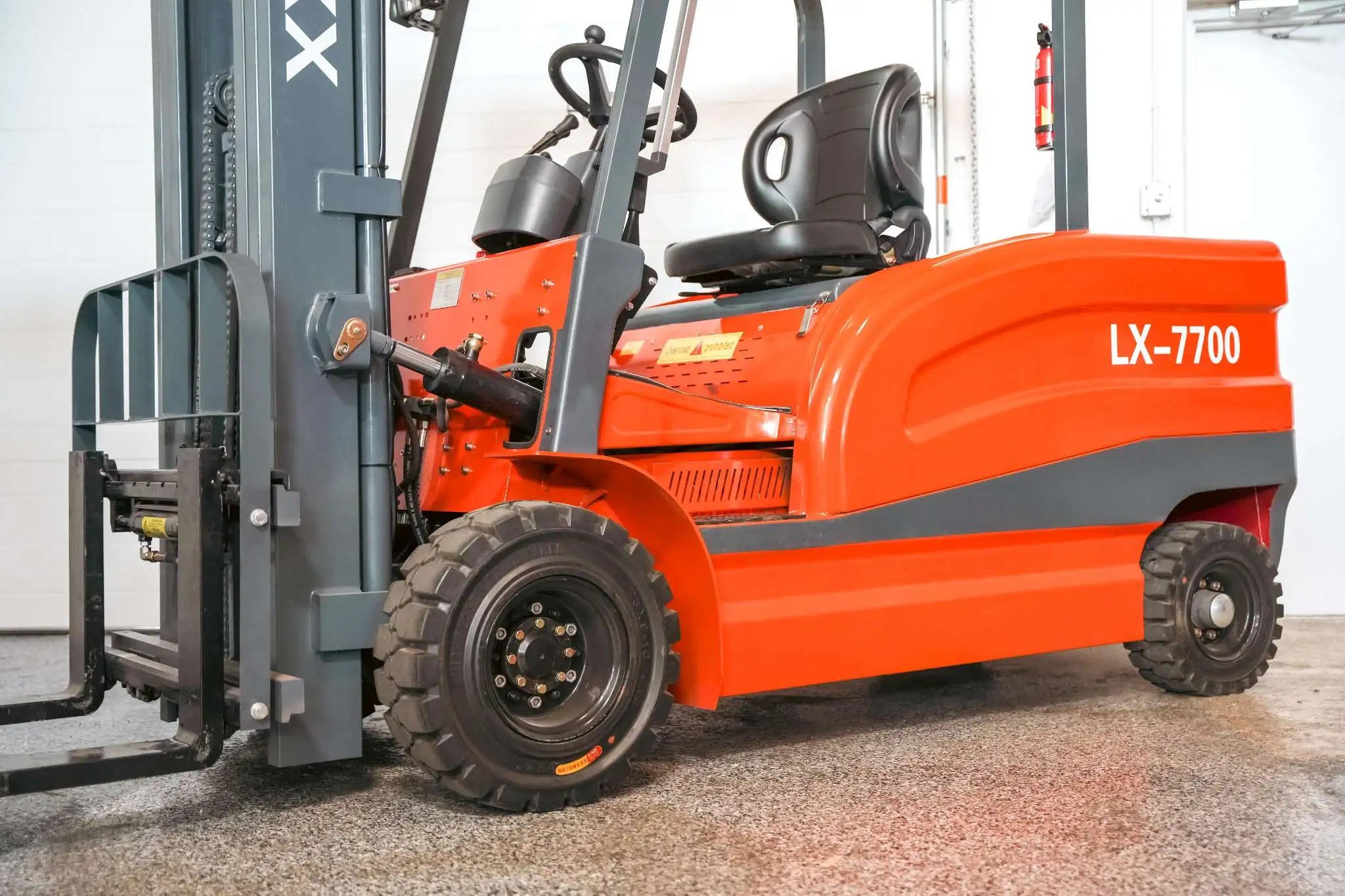 Low-angle side view of the LUXX LX-7700 forklift highlighting the heavy-duty solid tyres, hydraulic cylinder, load backrest, and bold red chassis in a warehouse setting.