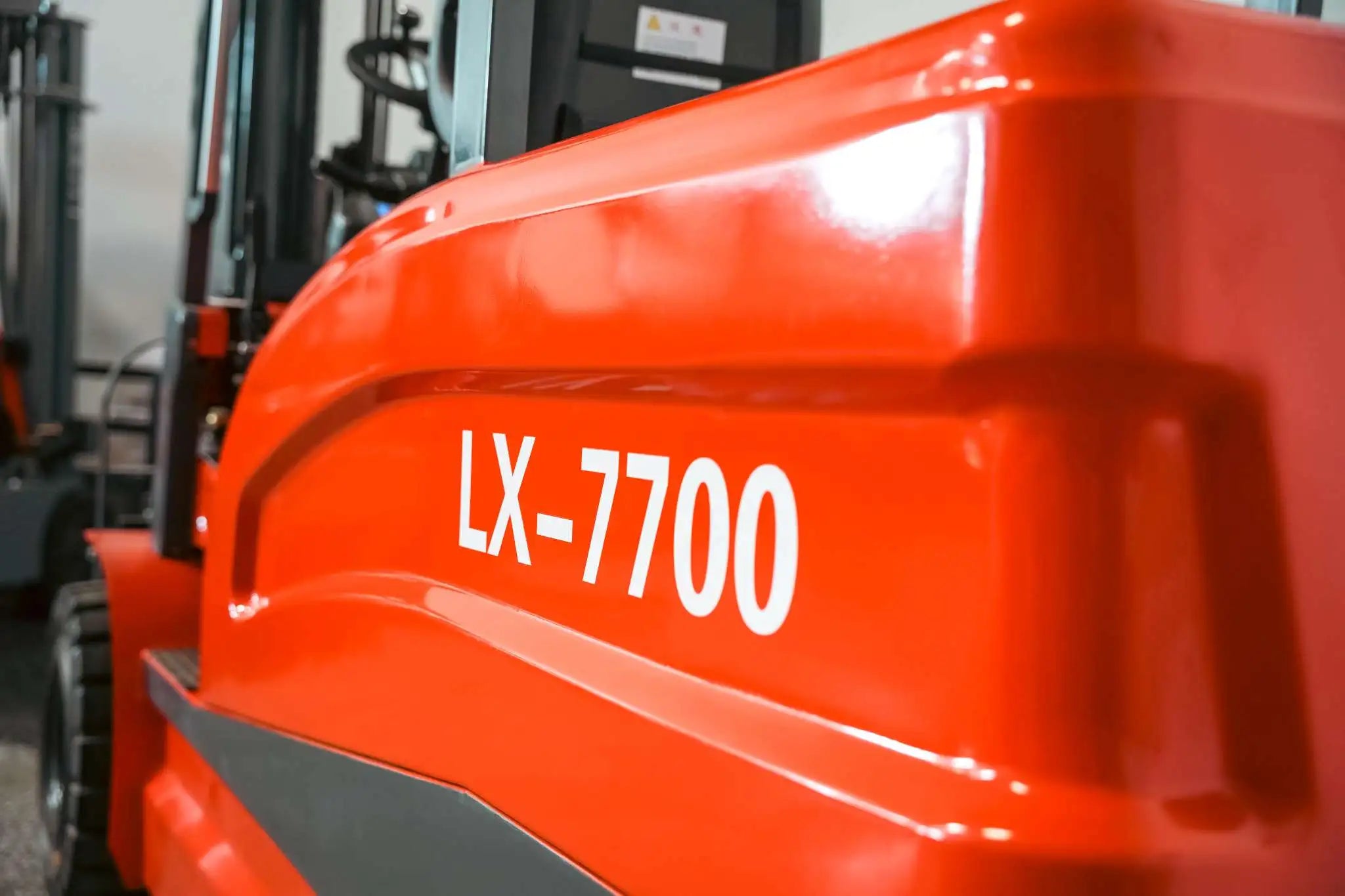 Close-up of the LUXX LX-7700 model badge on the glossy red forklift body panel, with a second forklift unit visible in the blurred background.