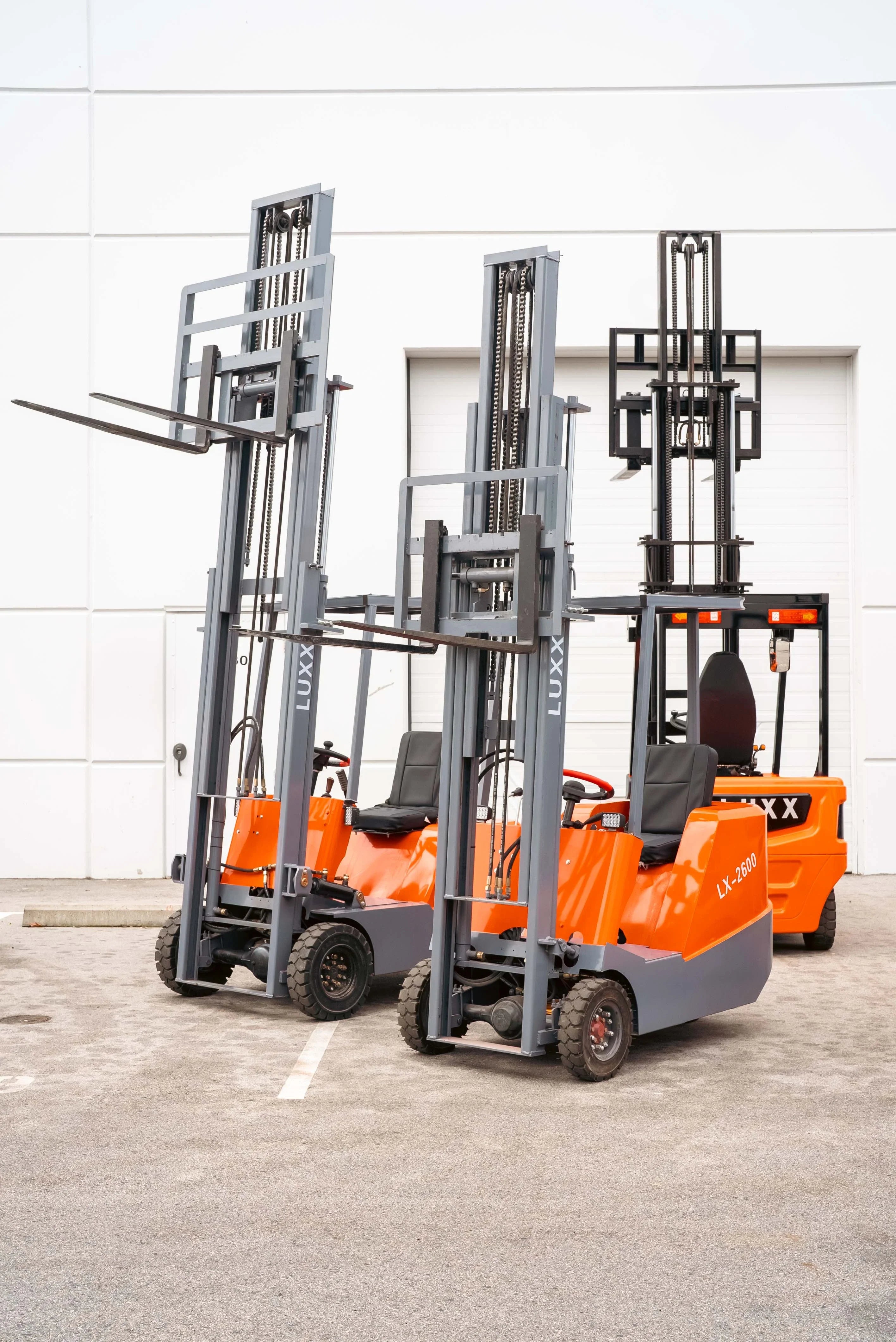Three LUXX electric forklifts including the LX-2600 parked outdoors with fully extended grey masts raised high, demonstrating lifting reach and compact orange body design.
