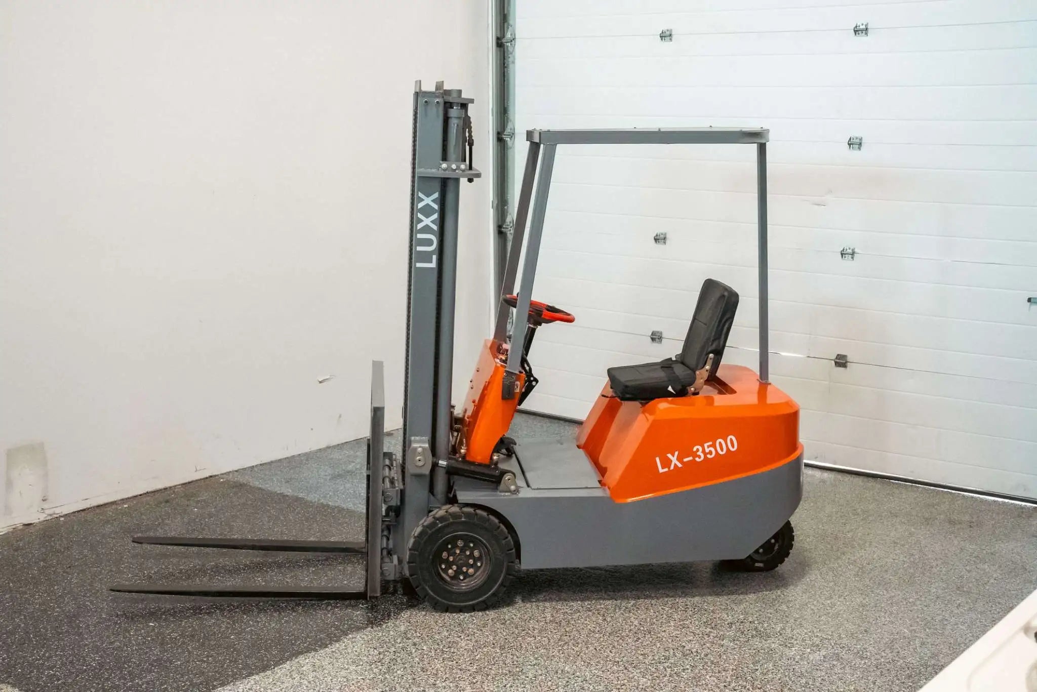 LUXX LX-3500 compact orange and grey forklift with single mast, open operator seat, red steering wheel, and small solid tyres, parked inside a clean warehouse facility.