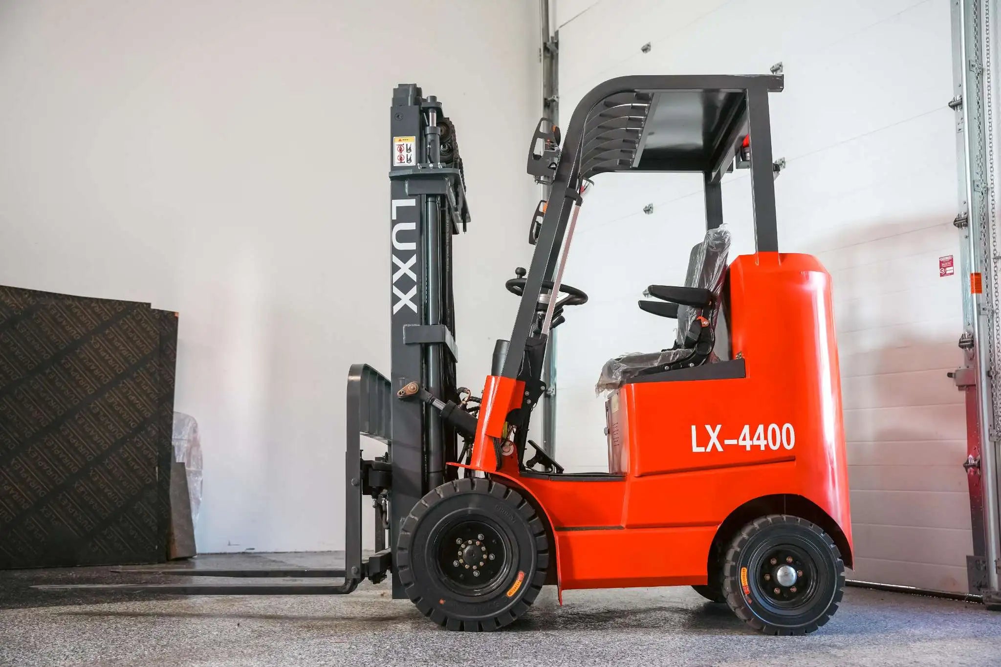 LUXX LX-4400 Compact red forklift with grey single-mast, enclosed overhead guard, still-wrapped seat, and solid tyres, positioned beside a warehouse garage door.