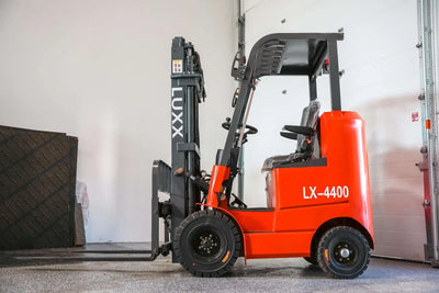 LUXX LX-4400 Compact red forklift with grey single-mast, enclosed overhead guard, still-wrapped seat, and solid tyres, positioned beside a warehouse garage door.