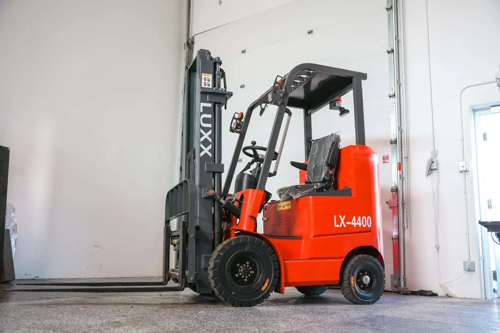 Low-angle front view of the LUXX LX-4400 Compact red forklift with grey mast, load backrest, enclosed overhead guard, and factory-wrapped seat beside a warehouse loading door.