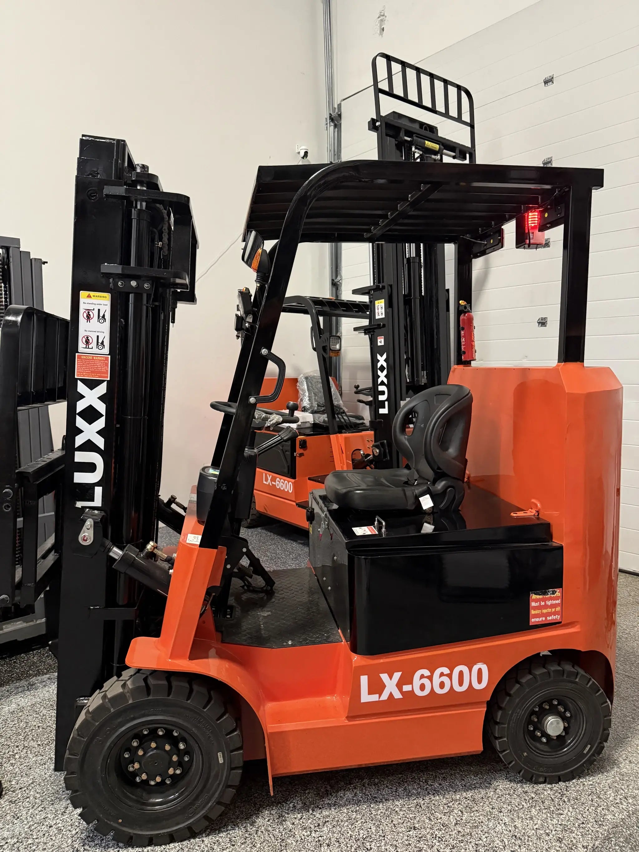 Rear three-quarter view of the LUXX LX-6600 Compact forklift revealing the ergonomic seat, red tail lights, overhead guard, and black mast in a multi-unit showroom.