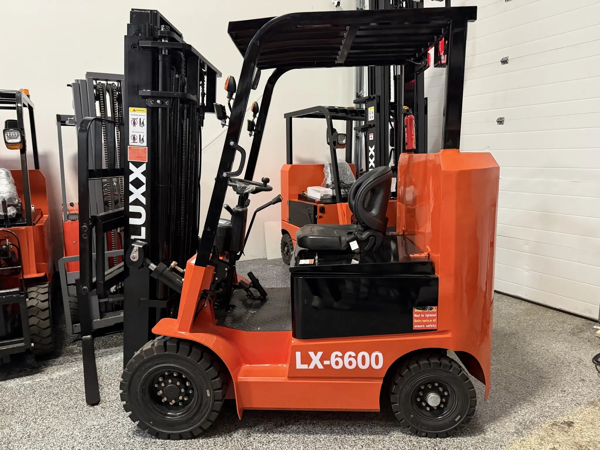 LUXX LX-6600 Compact orange forklift with black dual-mast, overhead guard, and solid tyres, displayed in a showroom alongside multiple other LUXX forklift models.
