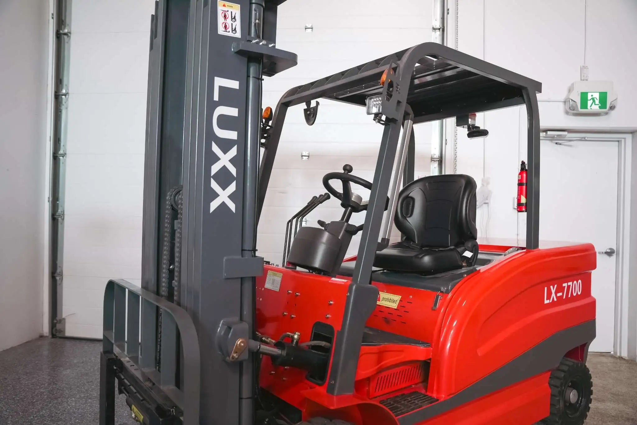 Close-up of the LUXX LX-7700 forklift operator cabin showing the steering wheel, ergonomic black seat, overhead guard, and grey mast with safety labels.
